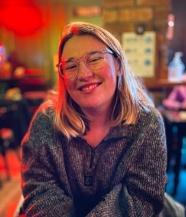 A woman with white skin, blond hair and glasses wearing a grey jumper smiling at the camera.